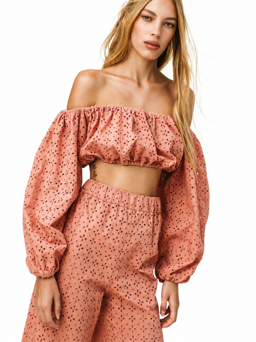 A woman modeling a matching two-piece set featuring an off-the-shoulder crop top and high-waisted trousers, both in a dusty rose pink color with a small black polka dot pattern, set against a plain white background.