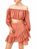 A woman wearing a terracotta-colored two-piece eyelet set consisting of an off-the-shoulder long-sleeve crop top and a matching ruffled mini skirt with a side bow, set against a plain white background.