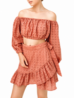A woman wearing a terracotta-colored two-piece eyelet set consisting of an off-the-shoulder long-sleeve crop top and a matching ruffled mini skirt with a side bow, set against a plain white background.