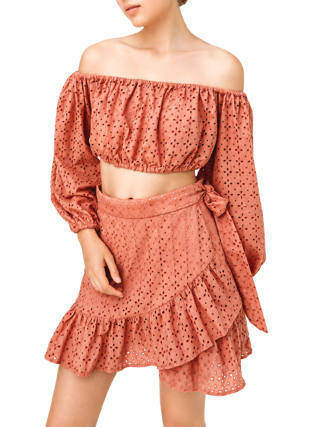 A woman wearing a terracotta-colored two-piece eyelet set consisting of an off-the-shoulder long-sleeve crop top and a matching ruffled mini skirt with a side bow, set against a plain white background.