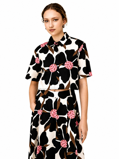 A model wearing a matching two-piece set consisting of a short-sleeve button-up shirt and a high-waisted skirt, both featuring a bold black and off-white floral pattern with pink flower centers and brown leaves, set against a plain white background.
