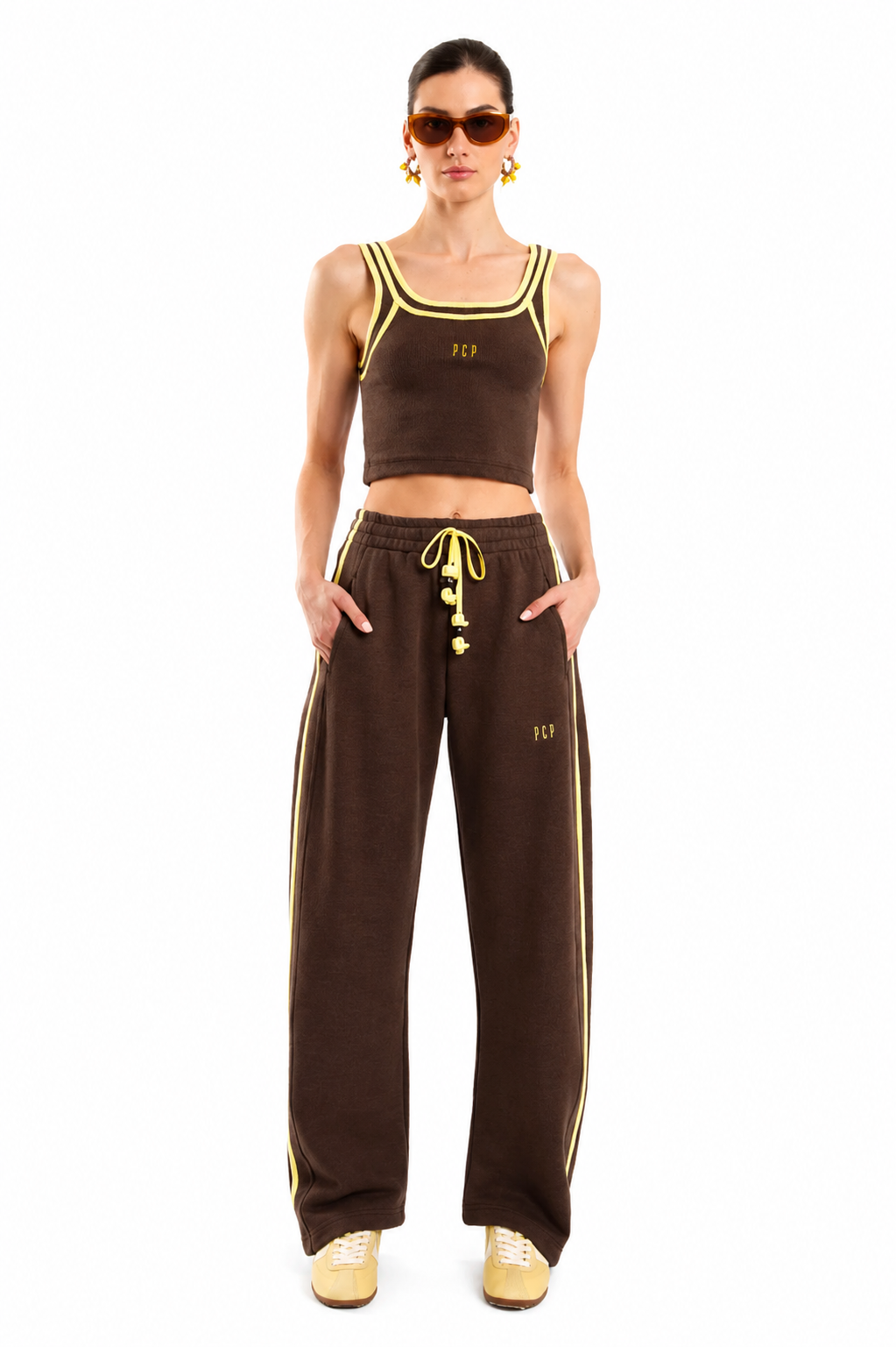 A model wearing a matching brown two-piece lounge set with yellow accents, including a cropped tank top and wide-leg sweatpants, against a plain white background.