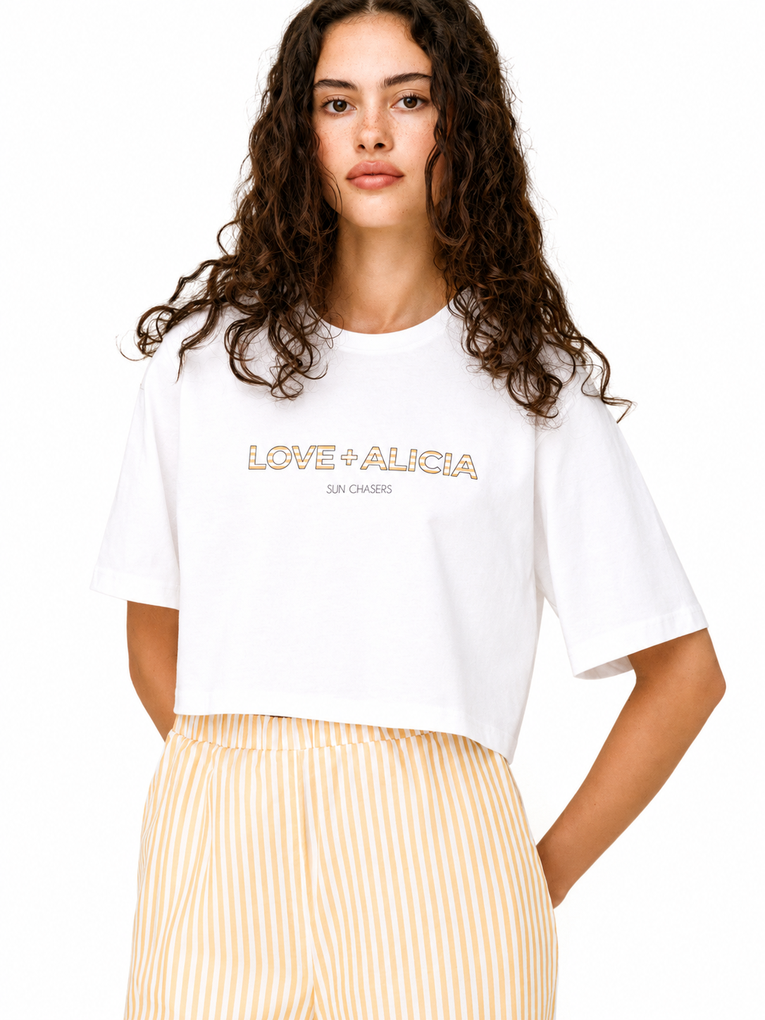 A woman modeling a white short-sleeve crop top with 'LOVE + ALICIA' printed in gold across the chest, paired with light yellow and white vertically striped pants, against a plain white background.