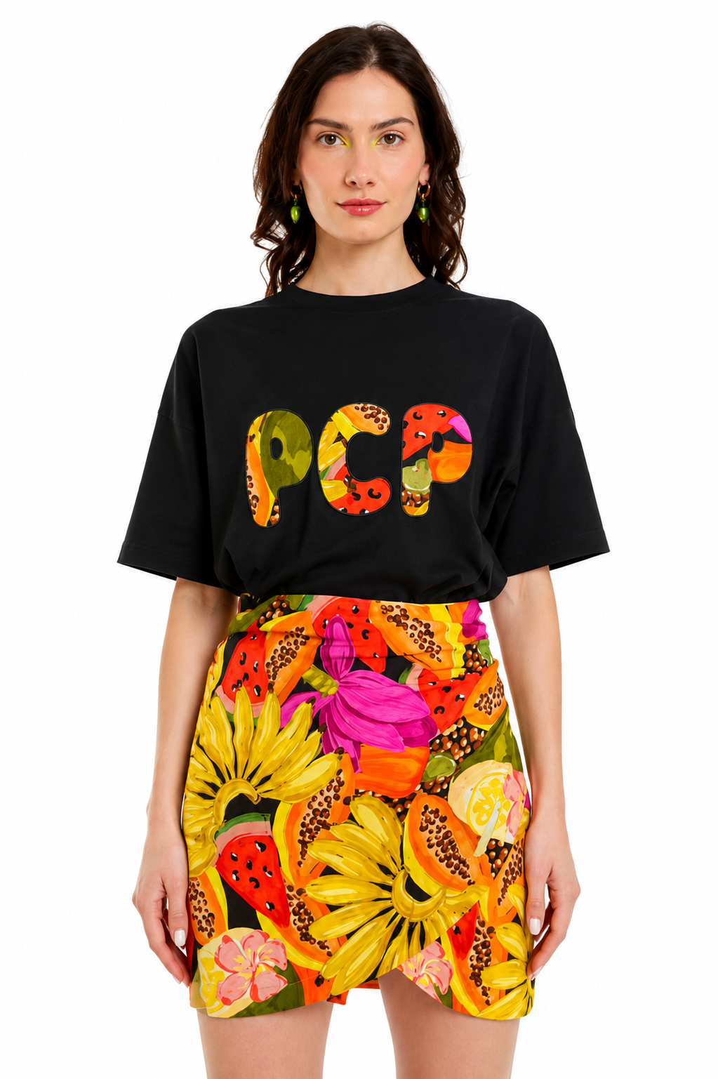 A model wearing a black oversized t-shirt with a colorful graphic print of the letters 'PCA' filled with fruit illustrations, paired with a vibrant yellow, orange, and pink floral print mini skirt, against a plain white background.