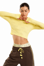 A woman modeling a pale yellow long-sleeve crop top with tonal repeating text, paired with brown sweatpants, against a plain white background.