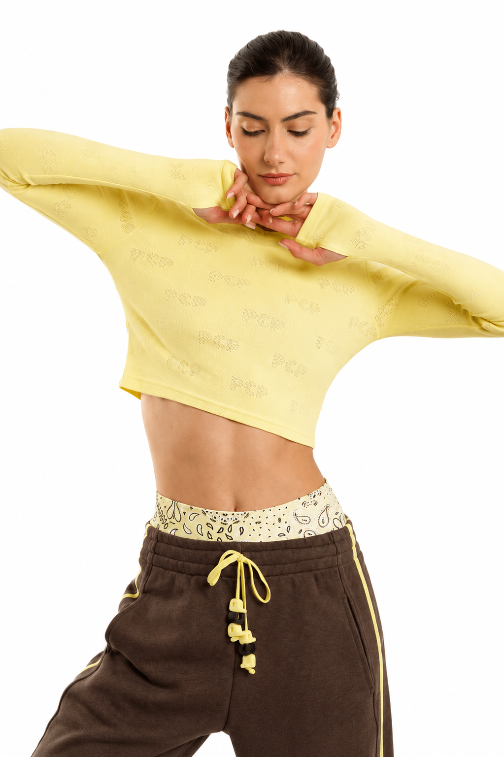 A woman modeling a pale yellow long-sleeve crop top with tonal repeating text, paired with brown sweatpants, against a plain white background.