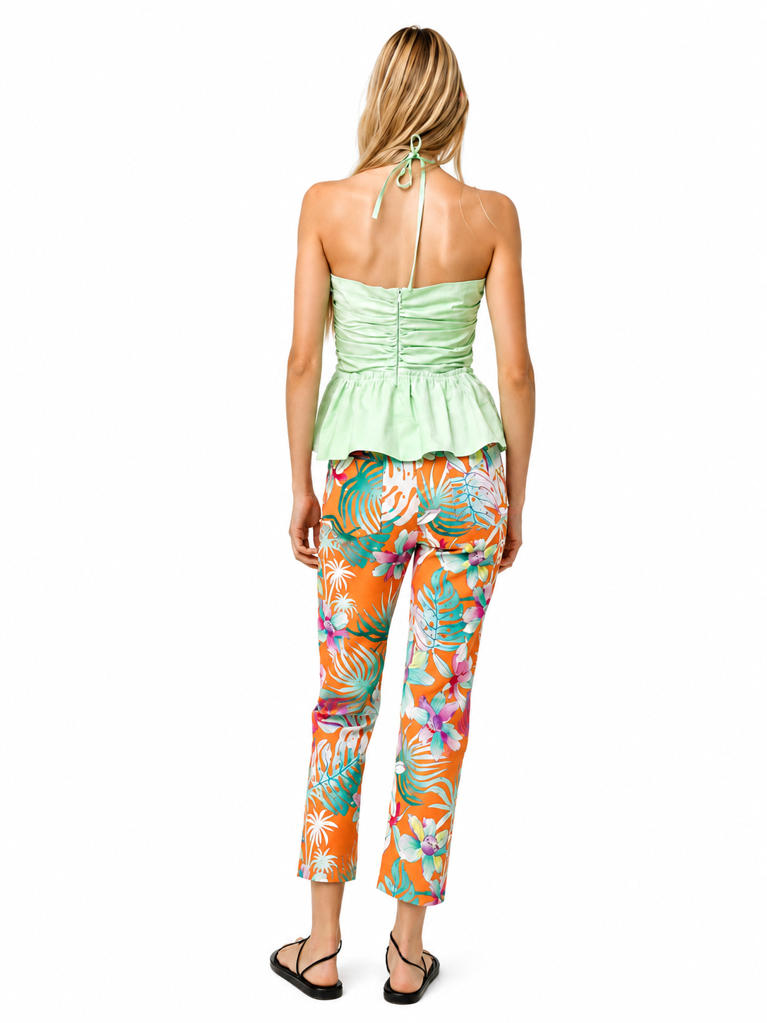 A woman shown from the back wearing a mint green strapless ruched peplum top and bright orange tropical floral print pants, paired with black sandals, against a plain white background.