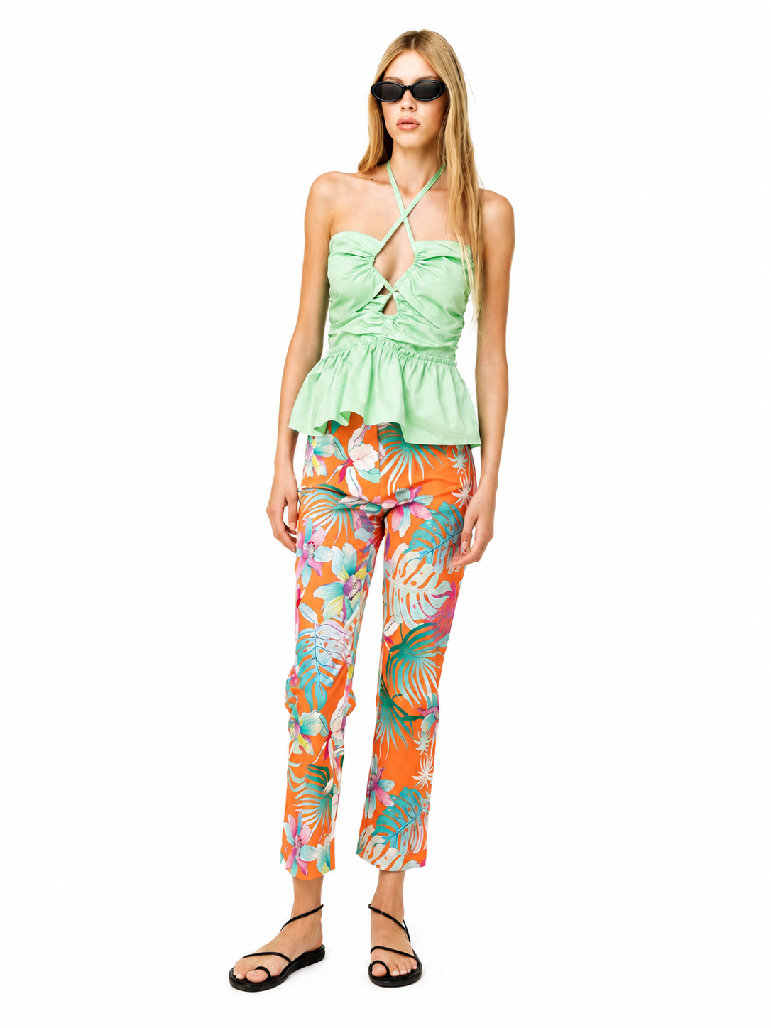 A woman modeling a light green halter crop top and bright orange tropical print pants against a plain white background.