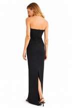A woman modeling a strapless black maxi dress from the back, featuring a fitted silhouette, a high slit, and black strappy heels against a plain white background.