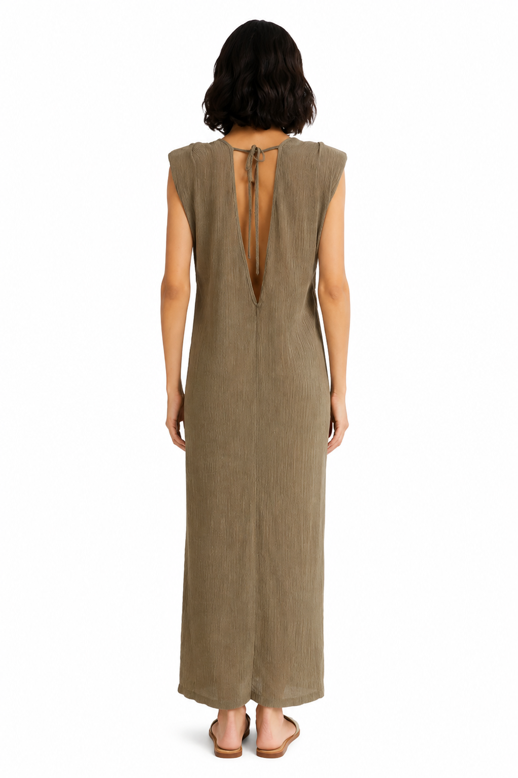 A woman viewed from the back wearing a sleeveless, floor-length taupe dress with a tie-back neckline, standing against a plain white background.