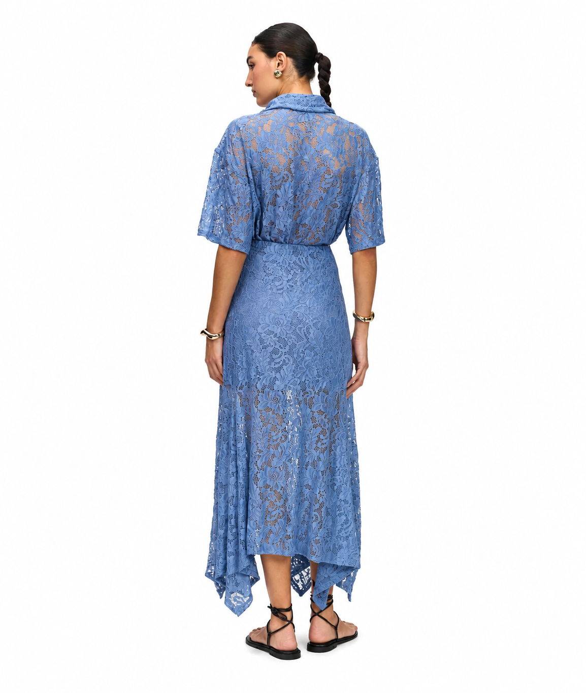 A woman viewed from the back wearing a blue lace midi dress with short sleeves, a collared neckline, and an asymmetrical draped hem, paired with black strappy sandals against a plain white background.