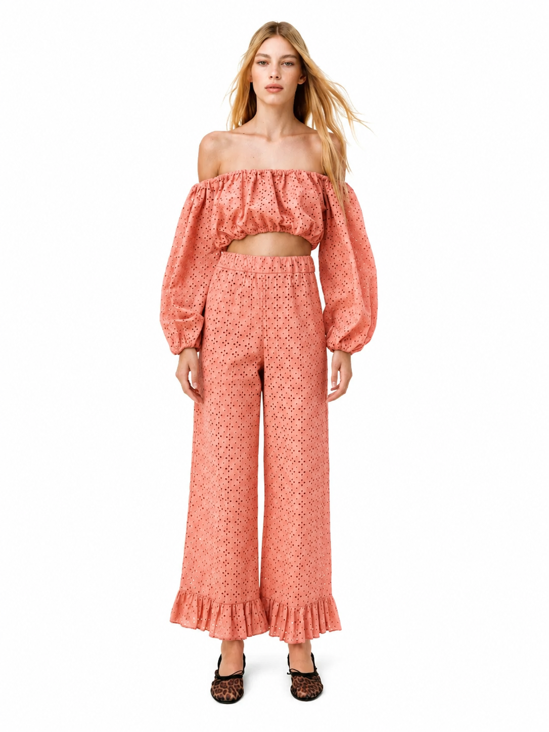A woman modeling a matching two-piece outfit consisting of an off-the-shoulder long-sleeve crop top and high-waisted wide-leg pants, both in a textured coral pink fabric with a small geometric pattern.