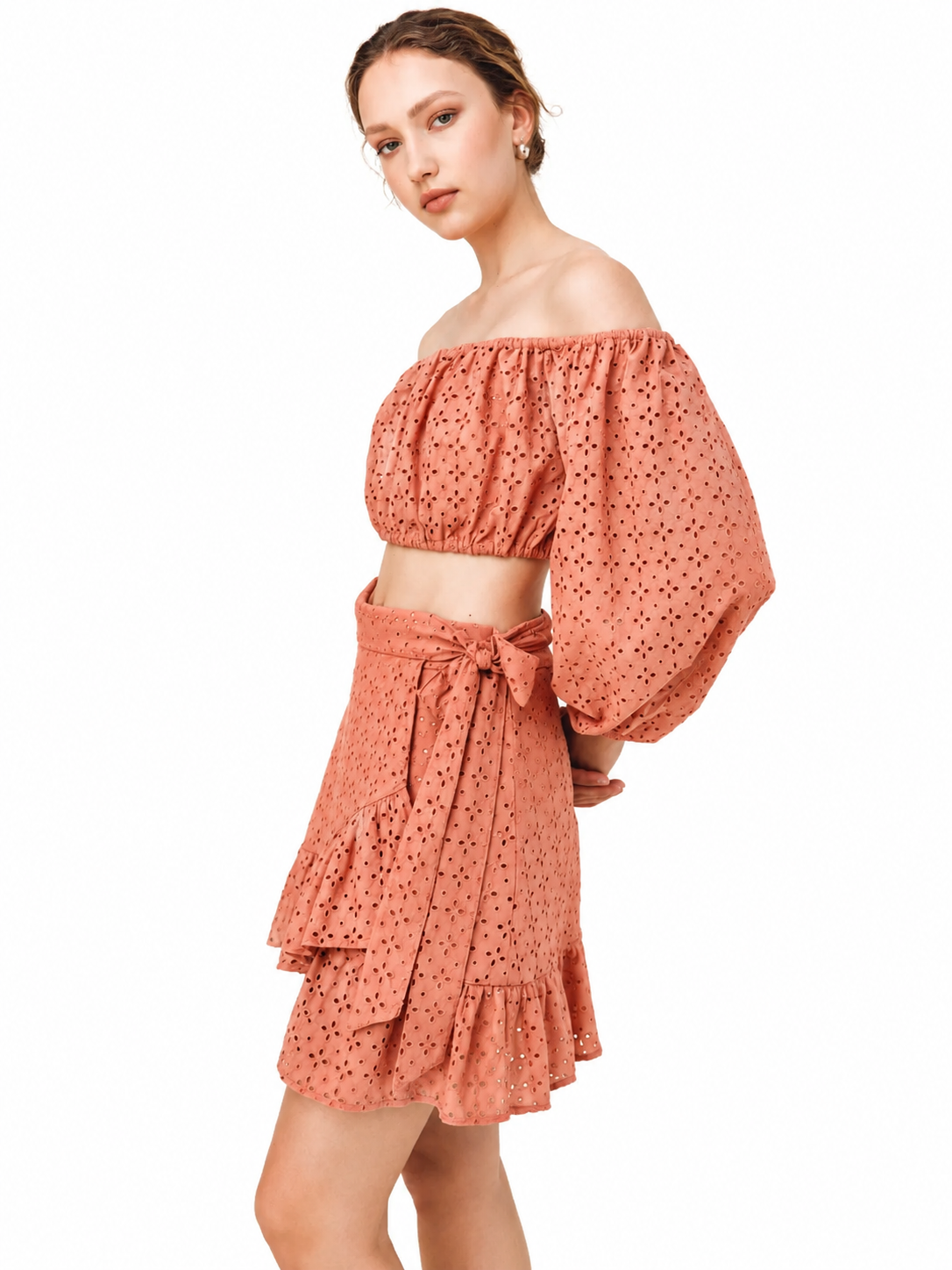 A woman modeling a terracotta orange off-the-shoulder crop top and matching tiered ruffle mini skirt with a small black dot pattern, set against a plain white background.