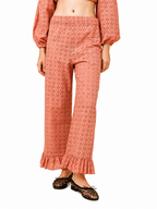 A model wearing a matching terracotta-colored eyelet two-piece set consisting of a cropped long-sleeve top and wide-leg ruffled pants, paired with leopard print flats, against a plain white background.