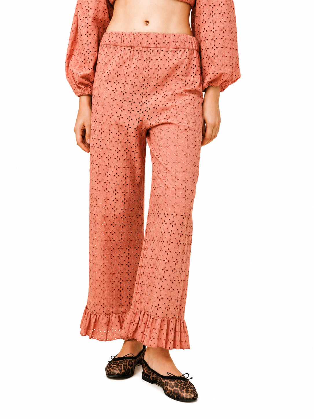 A model wearing a matching terracotta-colored eyelet two-piece set consisting of a cropped long-sleeve top and wide-leg ruffled pants, paired with leopard print flats, against a plain white background.