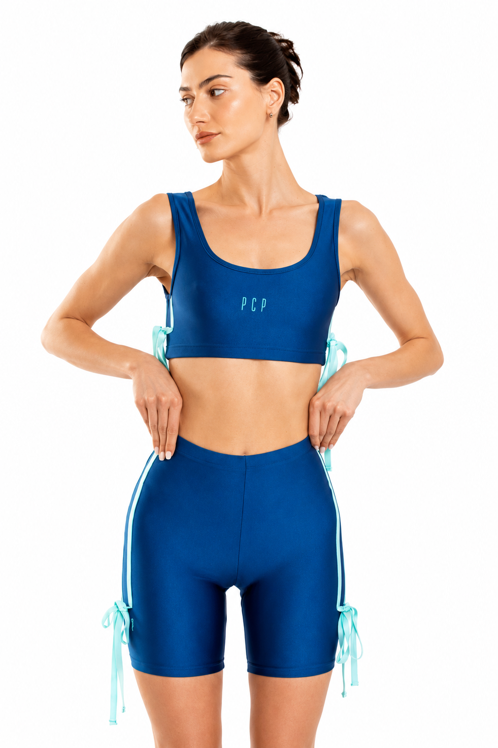 A woman modeling a matching royal blue activewear set consisting of a scoop-neck sports bra and high-waisted biker shorts with light blue side-tie details, set against a plain white background.