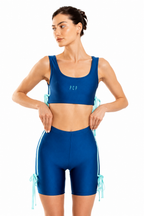 A woman modeling a matching royal blue activewear set consisting of a scoop-neck sports bra and high-waisted biker shorts with light blue side-tie details, set against a plain white background.