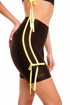 A close-up side view of a model wearing dark brown athletic biker shorts with bright yellow drawstring side details and a small gold 'PCP' logo near the hem, against a plain white background.