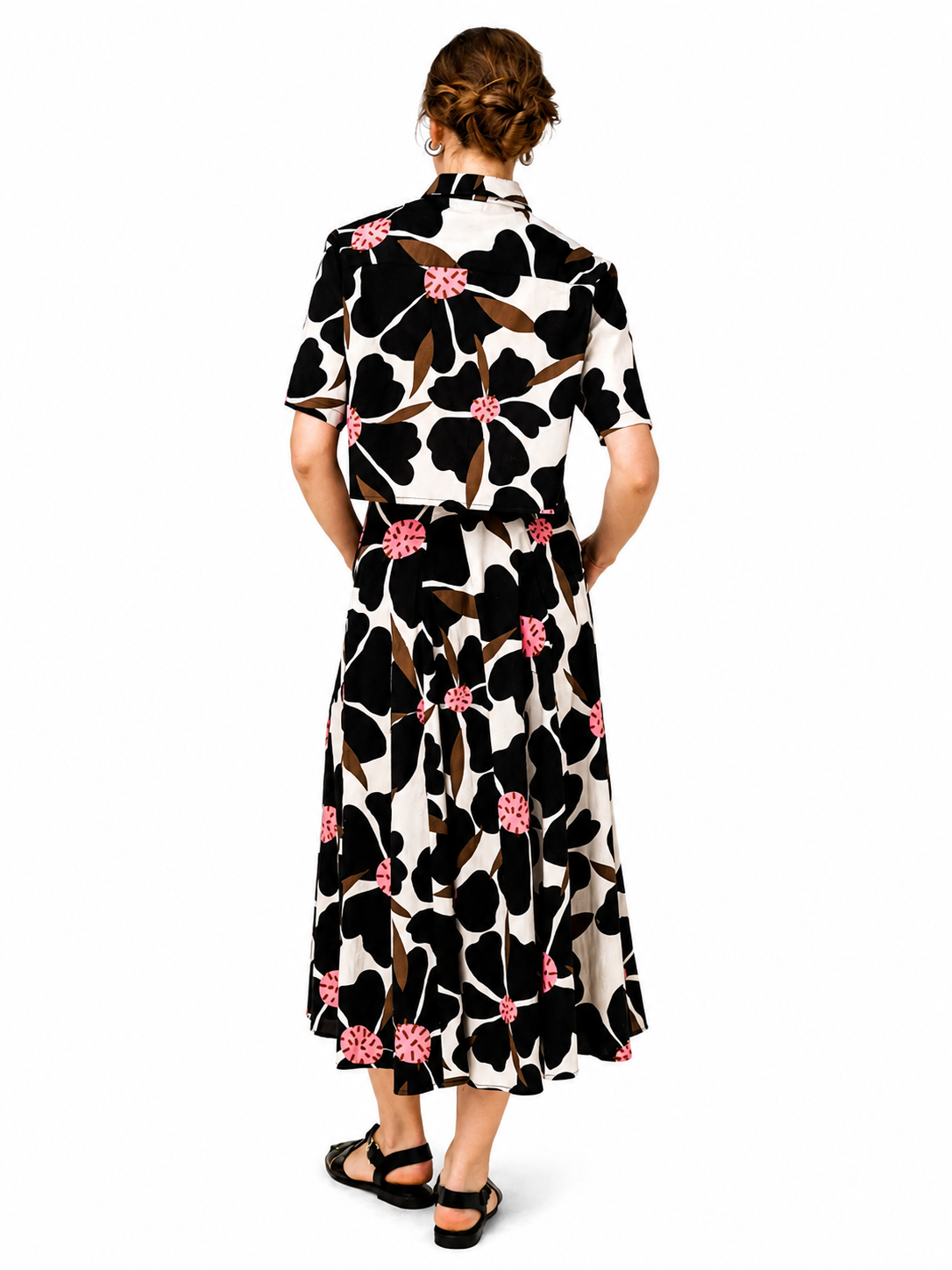 A woman viewed from behind wearing a short-sleeve collared midi dress with a bold black, white, and brown floral pattern accented by pink flower centers, paired with black sandals on a white background.