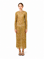 A woman modeling a long-sleeved midi dress with an all-over geometric floral pattern in mustard yellow and olive green, paired with black strappy sandals, against a plain white background.