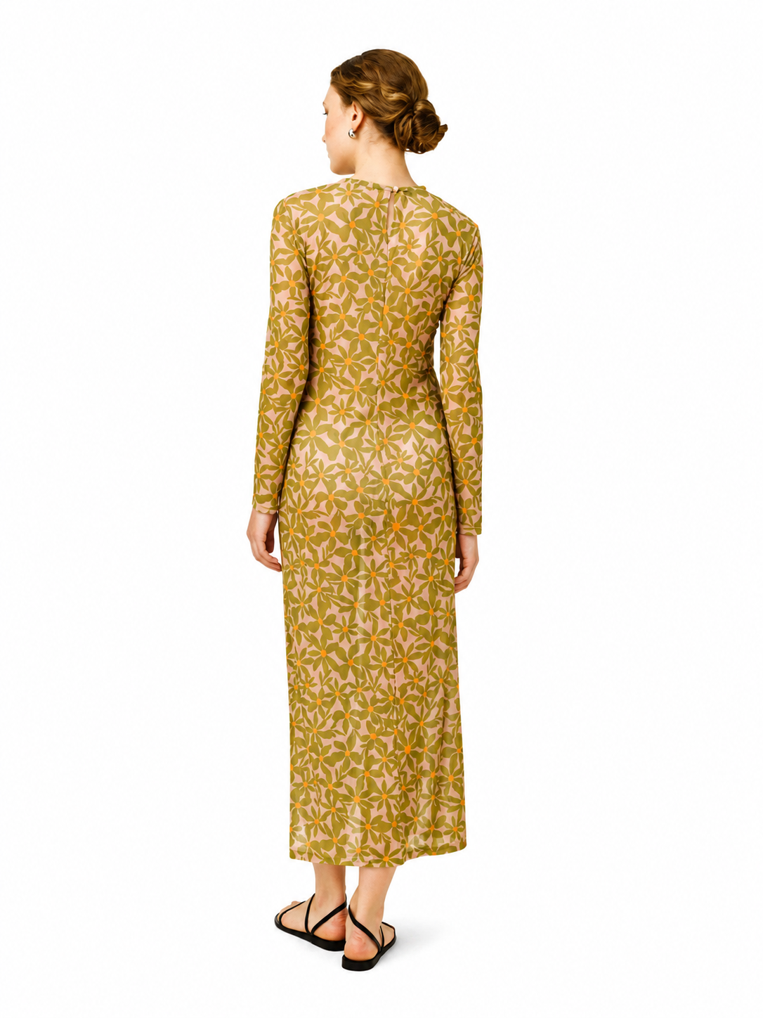 A woman viewed from the back wearing a long-sleeve midi dress with a dense yellow and green floral pattern on a beige base, paired with black strappy flat sandals, against a plain white background.
