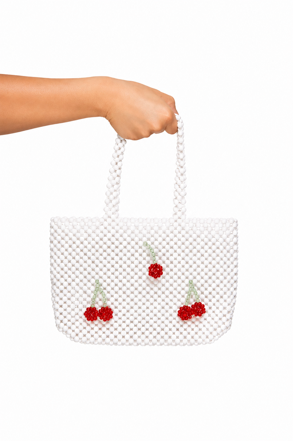 A white beaded tote bag with three red cherry embellishments, held by a person's hand against a solid white background.