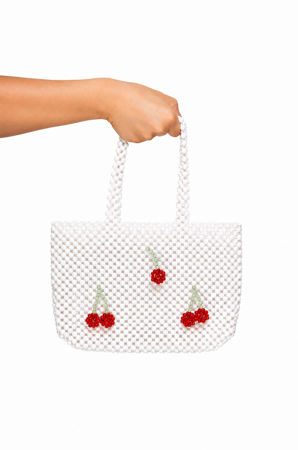 A white beaded tote bag with three red cherry embellishments, held by a person's hand against a solid white background.