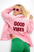 Person wearing a pink jacket with 'GOOD VIBES' text, green pants, and sunglasses on a white background