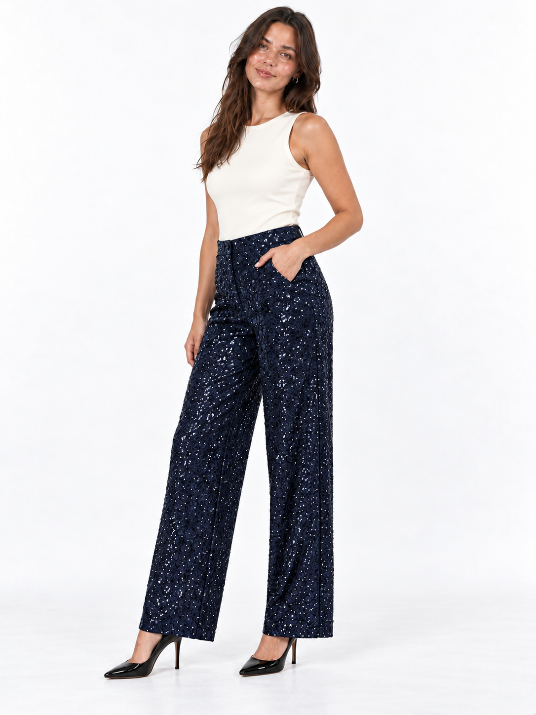 A woman modeling a pair of navy blue wide-leg trousers covered in small silver sequins, paired with a white sleeveless top and black heels, against a plain white studio background.