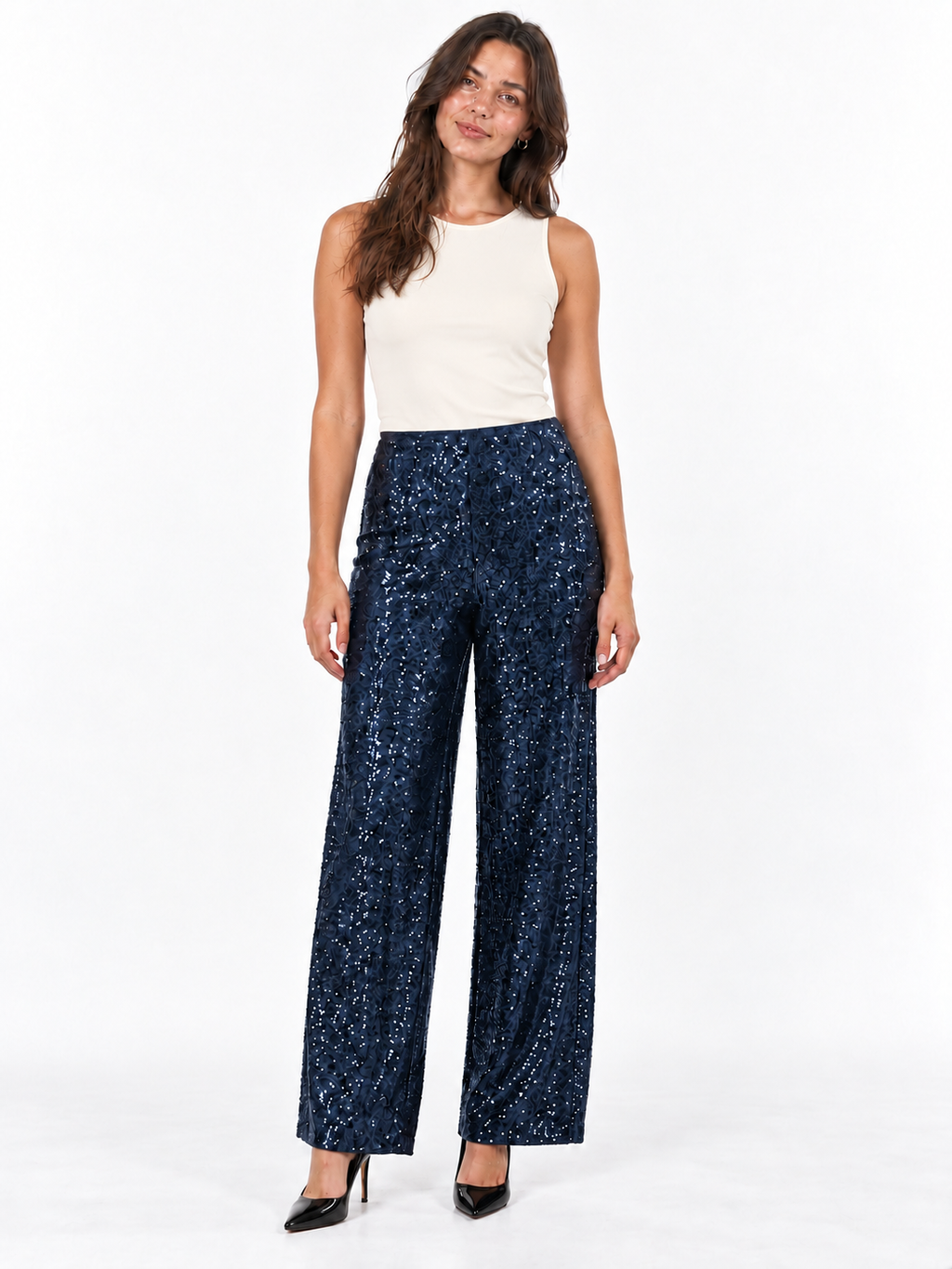 A woman modeling a pair of high-waisted, wide-leg navy blue pants covered in small gold sequins, paired with a cream sleeveless top and black heels, against a plain white studio background.
