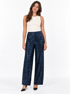 A woman modeling a pair of high-waisted, wide-leg navy blue pants covered in small gold sequins, paired with a cream sleeveless top and black heels, against a plain white studio background.