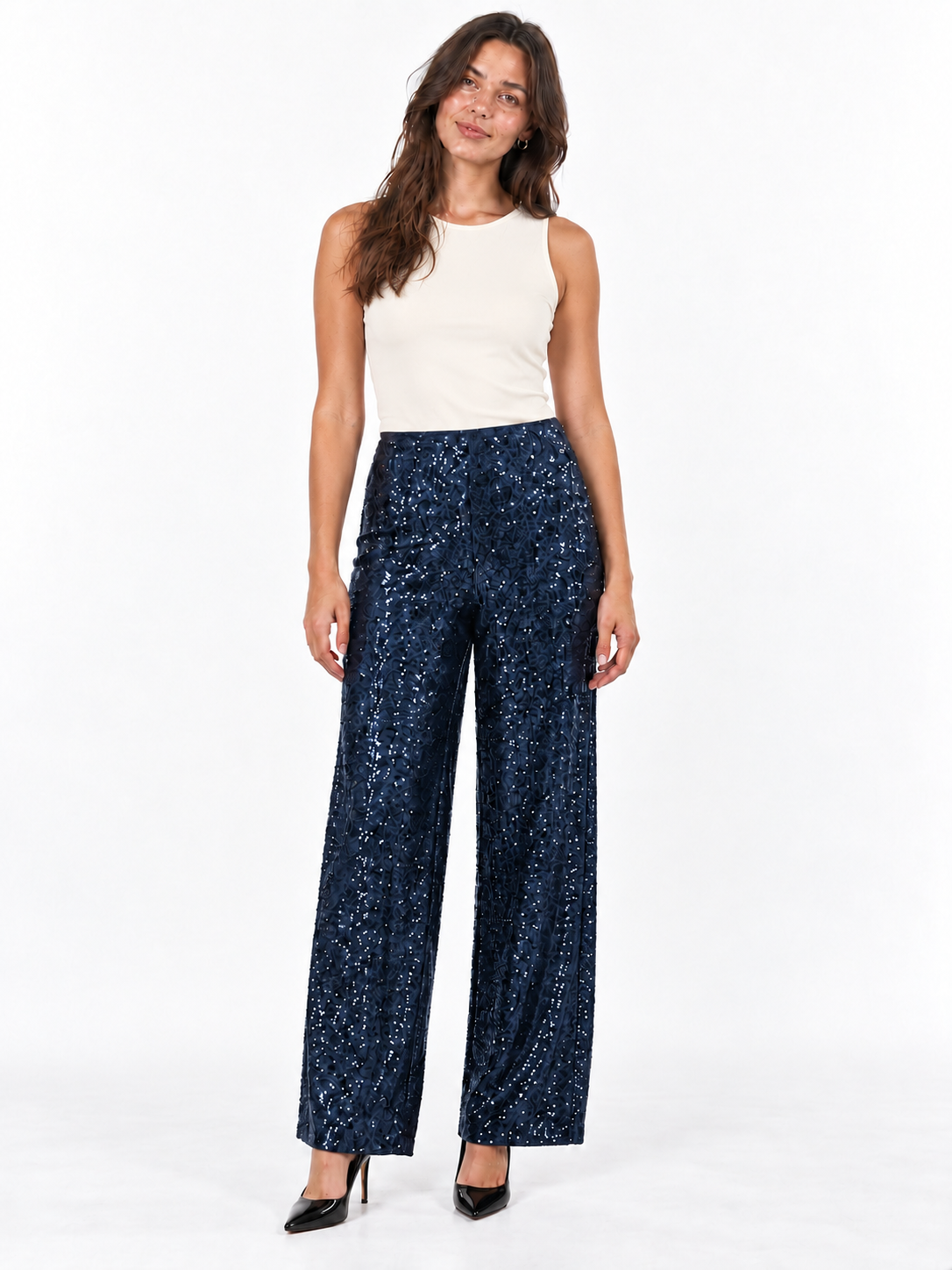 A woman modeling a pair of high-waisted, wide-leg navy blue pants covered in small gold sequins, paired with a cream sleeveless top and black heels, against a plain white studio background.