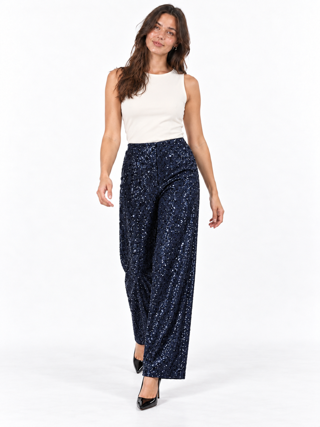 A woman modeling a pair of navy blue sequined wide-leg pants with a white sleeveless top and black heels, standing against a plain white studio background.