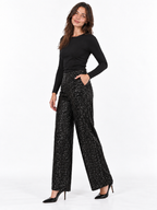 A woman modeling a pair of black wide-leg pants covered in small silver sequins, paired with a black long-sleeve top and black pointed-toe heels, against a plain white studio background.