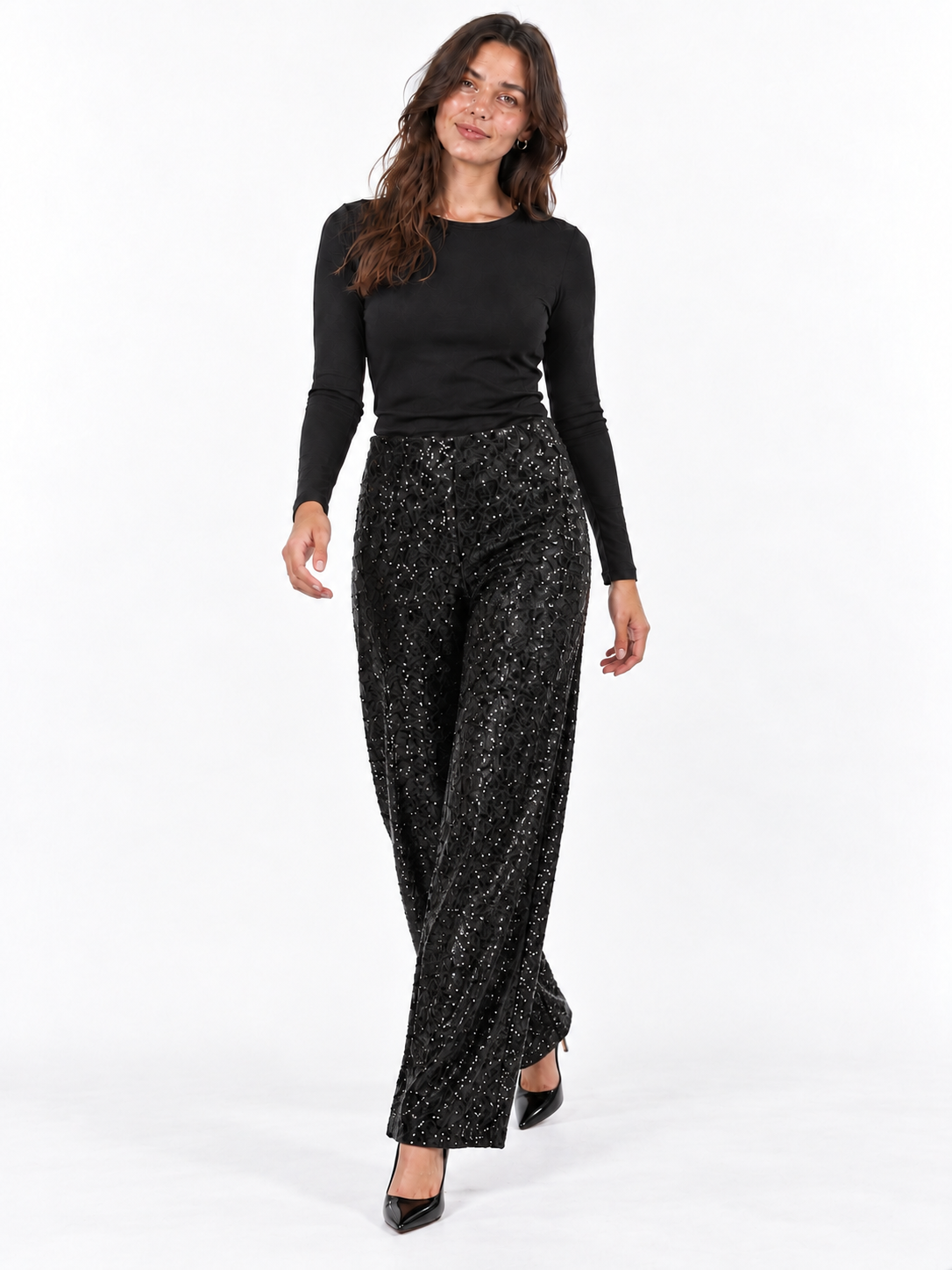 A woman modeling a black long-sleeve top and black wide-leg pants covered in dense sequins, paired with black pointed-toe heels, against a plain white studio background.