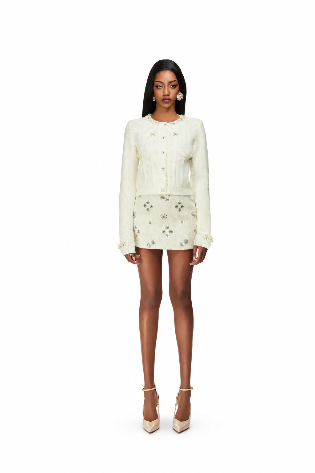 A model stands against a solid white background wearing a cream-colored two-piece outfit consisting of a long-sleeve button-front jacket and a matching mini skirt with small floral embroidery. She is wearing beige platform high heels.