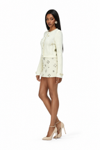 A woman modeling a cream-colored two-piece set consisting of a long-sleeve button-front jacket and a matching mini skirt with silver floral embroidery, paired with beige high-heeled sandals against a plain white background.