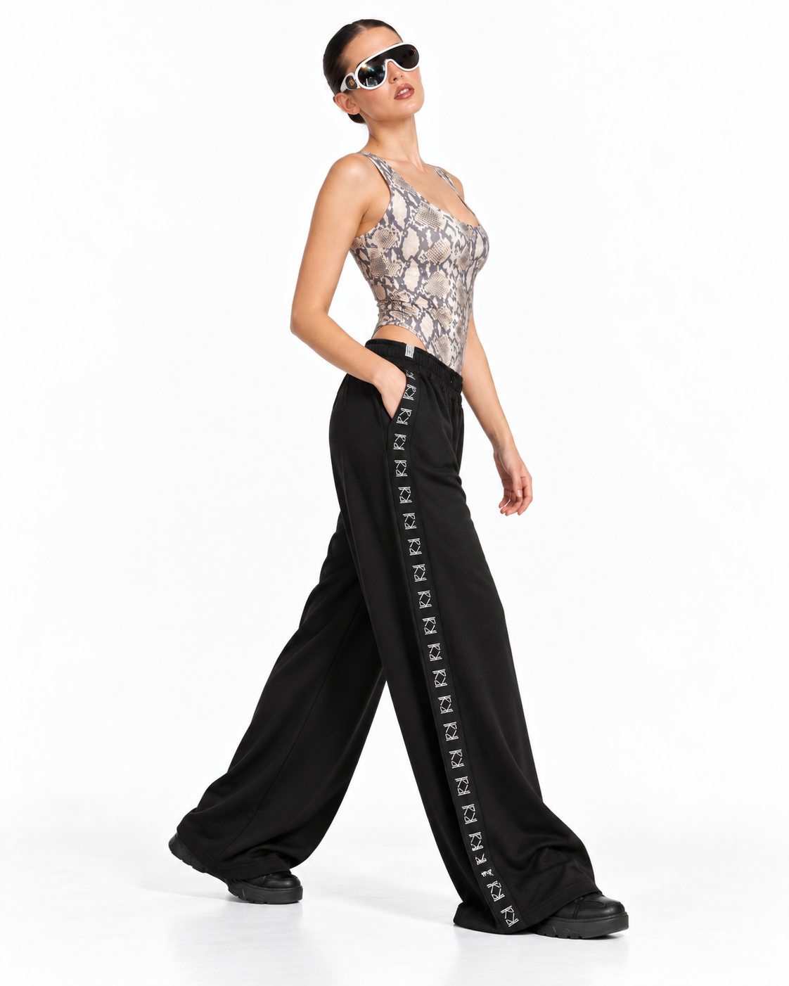 A model wearing a snakeskin-patterned bodysuit and black wide-leg trousers with a white geometric stripe detail, posing against a white studio background.