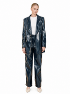 A model stands against a white studio background wearing a shiny dark navy blue faux leather pantsuit, consisting of a cropped blazer and matching wide-leg trousers, styled with a white ribbed top and beige pointed-toe heels.