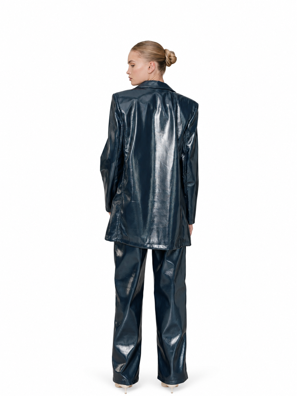 A model shown from the back wearing a shiny dark navy blue faux leather two-piece suit, consisting of a longline blazer and matching trousers, on a solid white background.