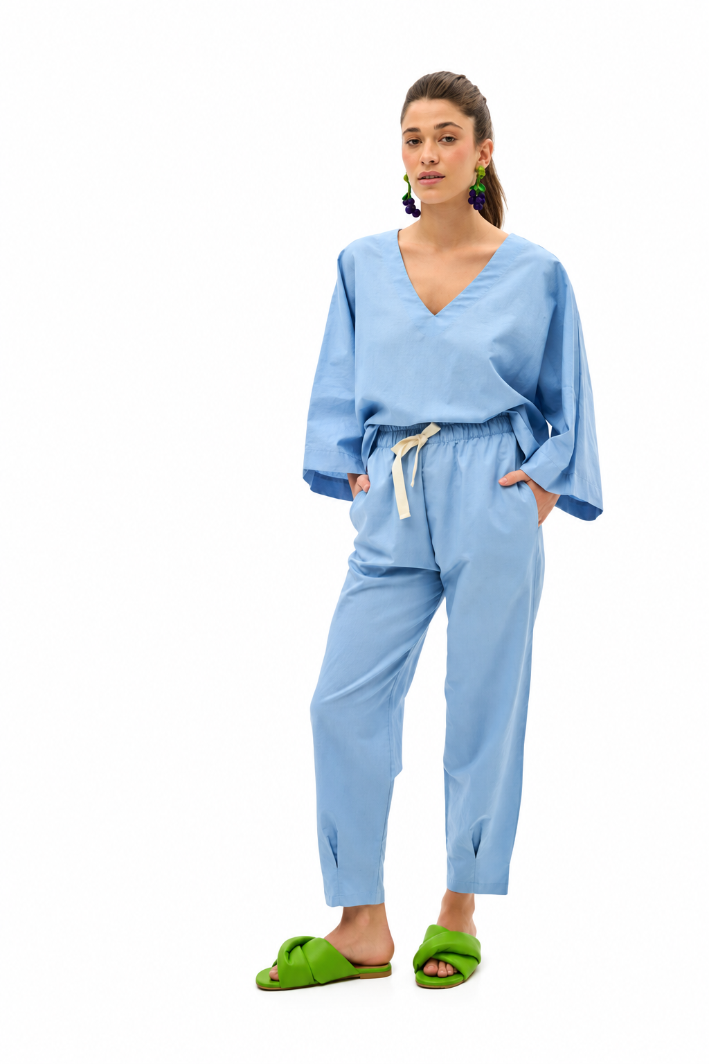 A woman modeling a light blue two-piece linen set consisting of a long-sleeve V-neck top and matching drawstring pants, paired with bright green slide sandals, on a solid white background.
