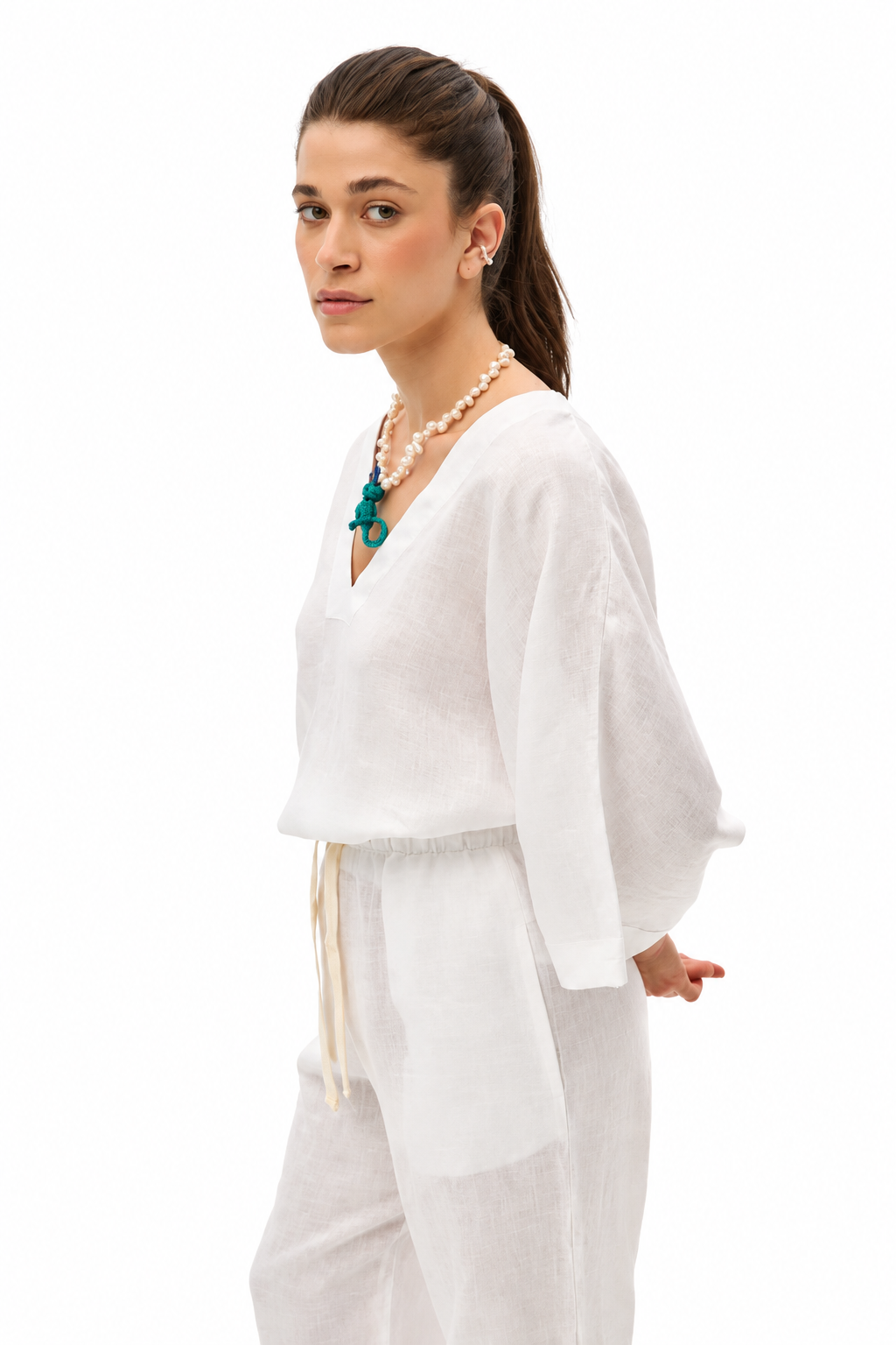 A woman modeling a white linen two-piece set consisting of a loose V-neck top with wide sleeves and matching wide-leg pants, styled with a pearl necklace and a green pendant against a solid white background.