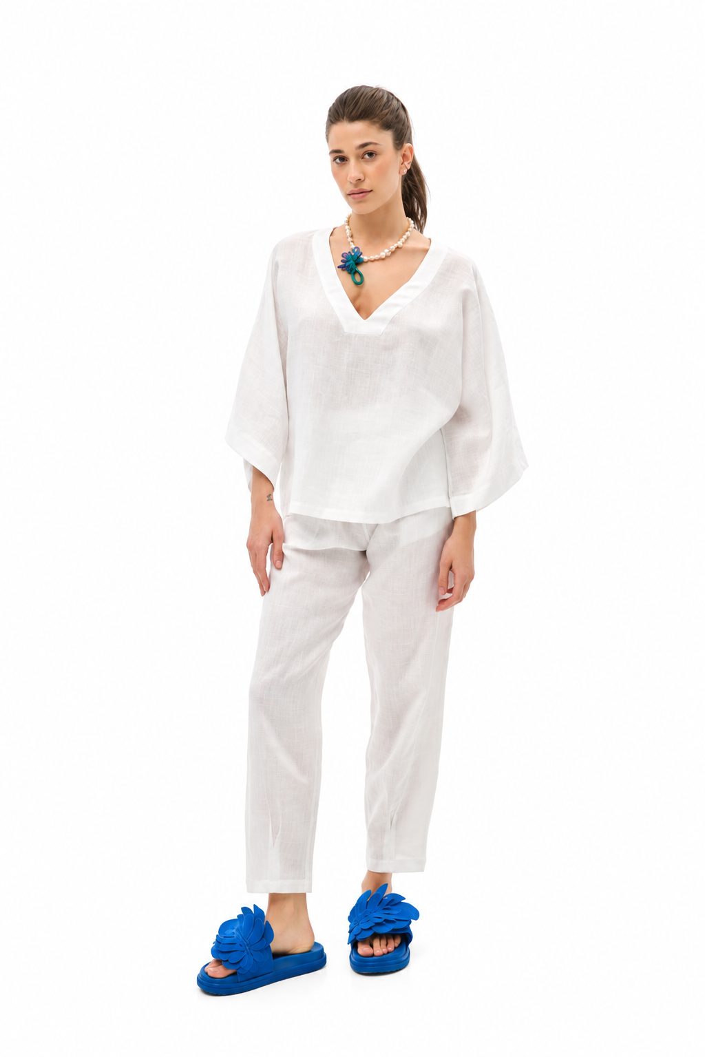 A woman modeling a white two-piece lounge set consisting of a loose, short-sleeved V-neck top and matching cropped pants, paired with bright blue slide sandals and a turquoise necklace, against a plain white background.