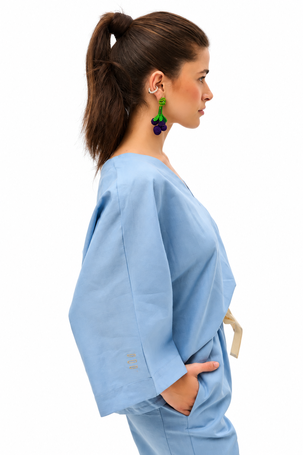 A woman in profile wearing a light blue scrub top and matching pants, with a high ponytail and colorful grape-shaped earrings, against a plain white background.