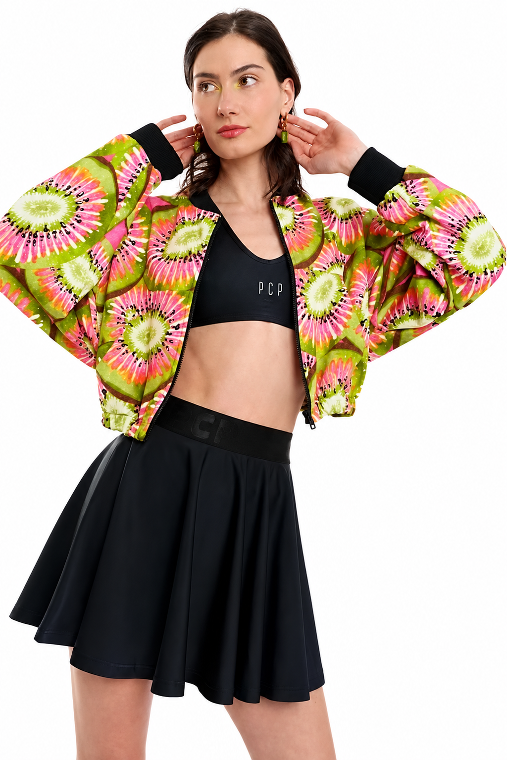 A model wearing a vibrant, cropped bomber jacket with a bright pink, green, and yellow kiwi-pattern print, styled over a black sports bra and a black pleated tennis skirt against a solid white background.