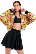 A model wearing a vibrant, cropped bomber jacket with a bright pink, green, and yellow kiwi-pattern print, styled over a black sports bra and a black pleated tennis skirt against a solid white background.