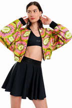 A model wearing a vibrant, cropped bomber jacket with a bright pink, green, and yellow kiwi-pattern print, styled over a black sports bra and a black pleated tennis skirt against a solid white background.