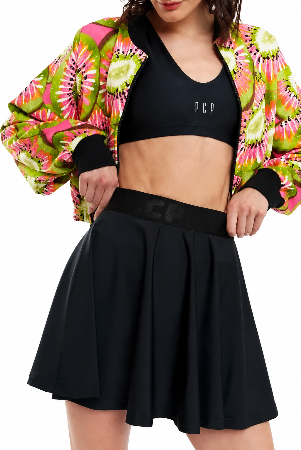 A model wearing a black athletic crop top with 'PCP' logo, a black pleated mini skirt with a matching black waistband, and a vibrant pink, green, and yellow floral bomber jacket, set against a plain white background.