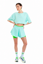 A model wearing a mint green cropped t-shirt and matching athletic shorts with neon green trim, paired with white socks and athletic sneakers, on a solid white background.