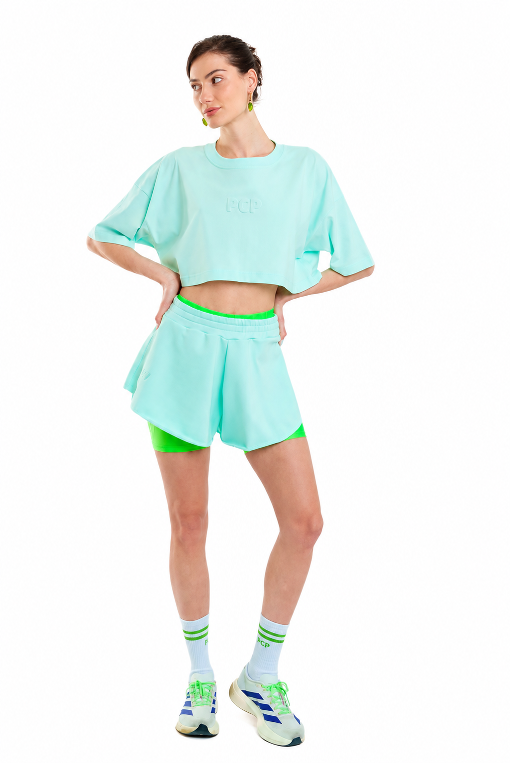 A model wearing a mint green cropped t-shirt and matching athletic shorts with neon green trim, paired with white socks and athletic sneakers, on a solid white background.